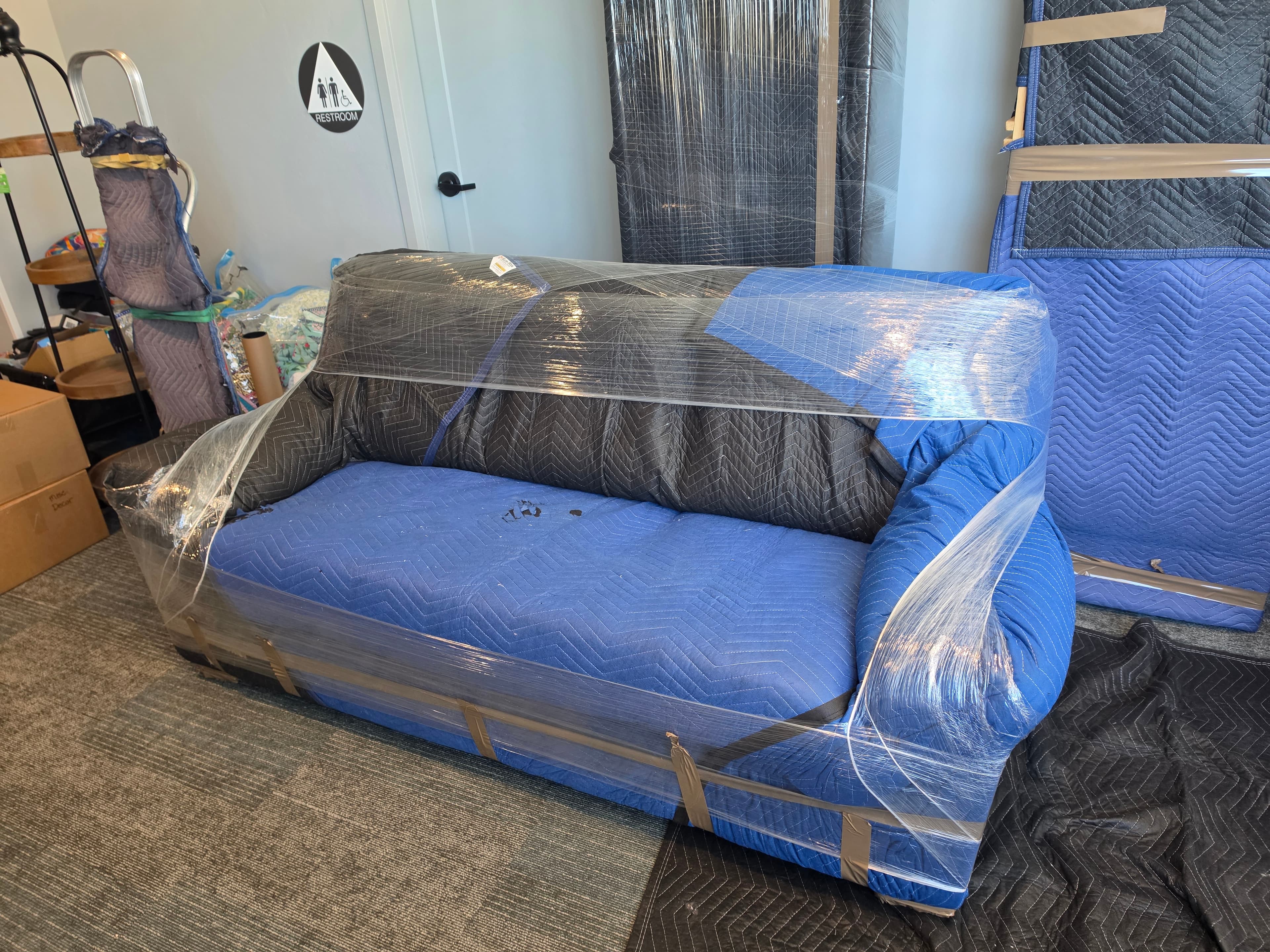 A couch double-wrapped in moving pads and shrink wrap, staged on floor protection